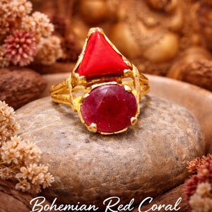 Peut inclure: Bague en or avec un triangle de corail rouge et une pierre ovale en rubis. La bague est posée sur une pierre avec le texte "Bohemian Red Coral & Ruby Gold Ring". Fond flou aux tons dorés.
