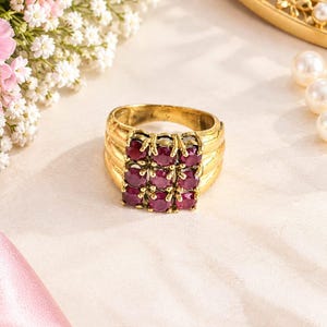 May include: A gold ring featuring a square cluster of nine round, deep red gemstones arranged in a three-by-three grid. The band of the ring is wide and constructed from three parallel gold bands that merge together at the base of the setting.