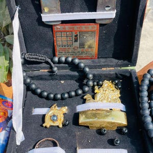 Op de afbeelding: Een open houten kist met diverse religieuze items. Inclusief een hoefijzer, een klein gouden beeld, een zwarte kralenarmband, een schildpendant, een ring en een kleine kaart met tekst. De items zijn gerangschikt op een zwart fluwelen oppervlak.
