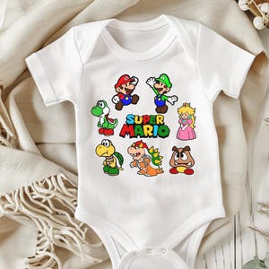 May include: White baby onesie featuring a colorful Super Mario graphic. The design includes Mario, Luigi, Princess Peach, and other characters from the video game series. The words "Super Mario" are also printed on the onesie.