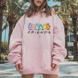 May include: A light pink hoodie with a front pocket and a drawstring hood. The front of the hoodie features a graphic of colorful Care Bears above the word "FRIENDS" in white block letters. The hoodie is made of a soft material.