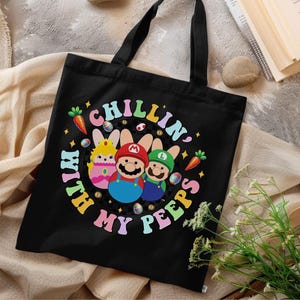 May include: Black tote bag featuring a colorful graphic of three cartoon characters dressed as Easter bunnies, with the text "CHILLIN' WITH MY PEEPS" in a circular design. The design includes carrots, eggs, and stars. The bag has two black handles.