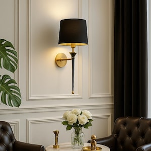 Könnte beinhalten: Eine Wandleuchte mit schwarzem Lampenschirm und goldenen Akzenten beleuchtet einen Raum. Ein kleiner runder Tisch mit Marmorplatte trägt eine Vase mit weißen Rosen. Zwei braune Ledersessel vervollständigen die Szene.