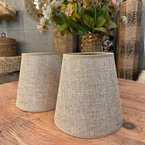Puede incluir: Dos pantallas de lámpara de tela beige con tejido texturizado, sobre una mesa de madera. Las pantallas tienen forma cónica y un color neutro, adecuadas para varios estilos de interior. El fondo incluye elementos decorativos y flores.