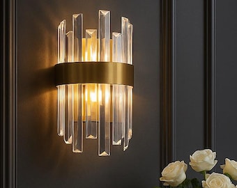 May include: A modern wall sconce with a gold-toned band and clear, rectangular crystal accents. The sconce is mounted on a dark wall, and the warm light it emits illuminates the surrounding area. A black marble-topped table with a bronze frame is below.