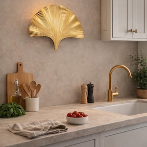 Könnte beinhalten: Eine goldfarbene, fächerförmige Wandleuchte ist an einer Wand in neutralen Tönen angebracht. Die Küchenszene umfasst einen Wasserhahn, Schränke und eine Arbeitsplatte mit einer Schüssel mit Tomaten, Holzutensilien und einem Schneidebrett.