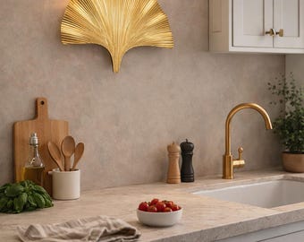 May include: A gold-coloured, fan-shaped wall sconce is mounted on a neutral-toned wall. The kitchen setting includes a faucet, cabinets, and countertop with a bowl of tomatoes, wooden utensils, and a cutting board.
