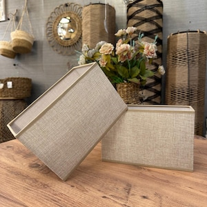 Puede incluir: Dos pantallas de lámpara rectangulares beige con tela texturizada se exhiben sobre una mesa de madera. Las pantallas tienen un color neutro y un diseño sencillo. El fondo incluye elementos decorativos como cestas y un espejo.
