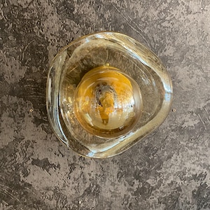 May include: An amber-toned glass light fitting with an unusual, organic form. It features a central bulb and an irregularly shaped glass shade. The fitting is mounted on a textured, dark grey wall.