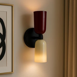 Könnte beinhalten: Moderne Wandleuchte mit schwarzem Sockel und zwei zylindrischen Schirmen. Der obere Schirm ist tiefrot, der untere cremefarben-gelb und strahlt ein warmes Licht aus. Die Leuchte ist an einer neutralfarbenen Wand montiert.