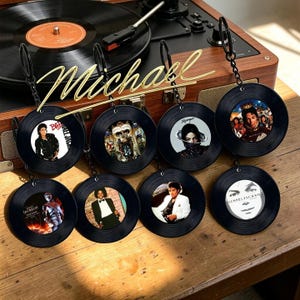 May include: Eight circular keychains designed to resemble miniature black vinyl records, each featuring a unique photographic portrait of Michael Jackson in the centre, attached to a black metal chain and a circular ring for attachment.