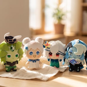 May include: Four plush dolls featuring stylized designs. The first is green with a dark hat, the second is white with blue accents, the third has white hair and a teal outfit, and the fourth is blue with a dark hood and a crescent moon emblem.
