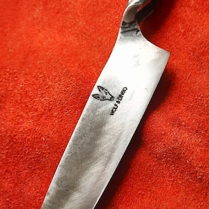 May include: A handcrafted chef's knife with a textured, silver-colored handle and a wide, polished blade. The blade features the text "WOLF & DINGO" and a logo. The knife is set against a red background.