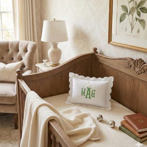 May include: A wooden cot with a white, scalloped-edge pillow embroidered with the letters "MAG" in green. A cream blanket is draped over the cot. A silver rattle and a stack of books are also visible.