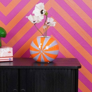 Può includere: Un mobile in legno nero con un telefono a disco verde, libri e un vaso decorativo con fiori. Il vaso ha strisce arancioni e grigie. Lo sfondo presenta un motivo a zig-zag rosa e arancione.