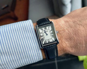 May include: A rectangular wristwatch with a silver case and a black leather band. The watch face is white with black Roman numerals and hands. The watch is on a wrist, with a blue and white striped shirt sleeve visible.