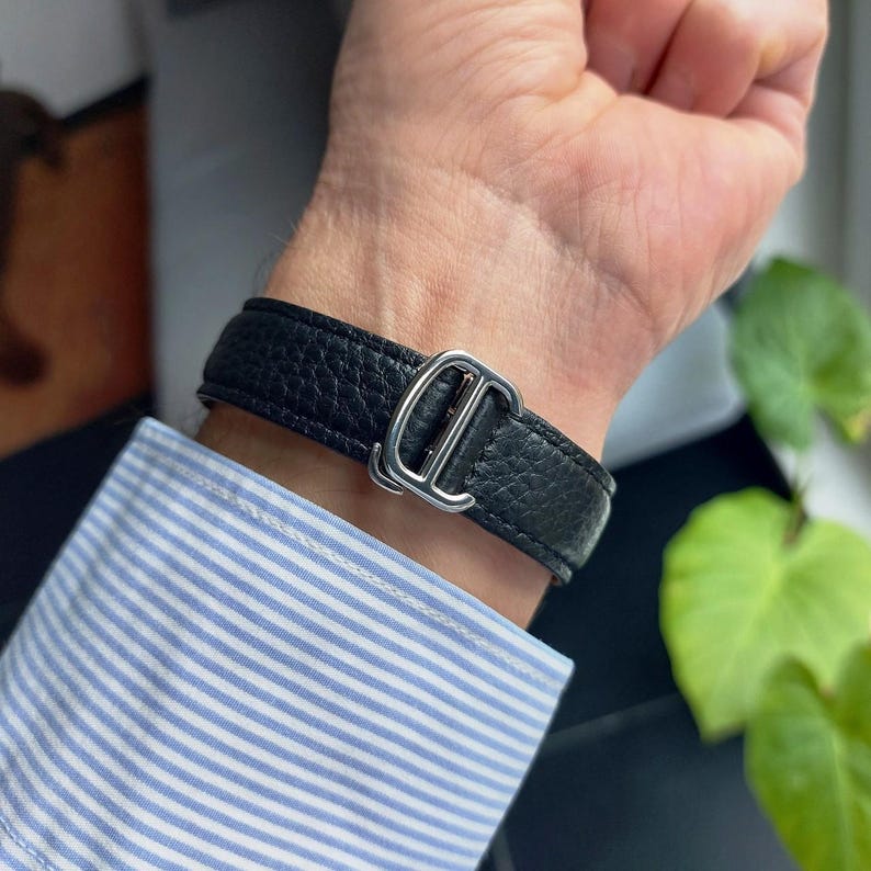 May include: Black leather bracelet with a silver-colored buckle. The bracelet is worn on a wrist, with a blue and white striped shirt sleeve visible in the background. The bracelet has a textured surface.
