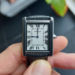 May include: A rectangular wristwatch with a silver-tone case and a black leather band. The watch face is white with black Roman numerals and the brand name "SANDA". The watch hands are blue.