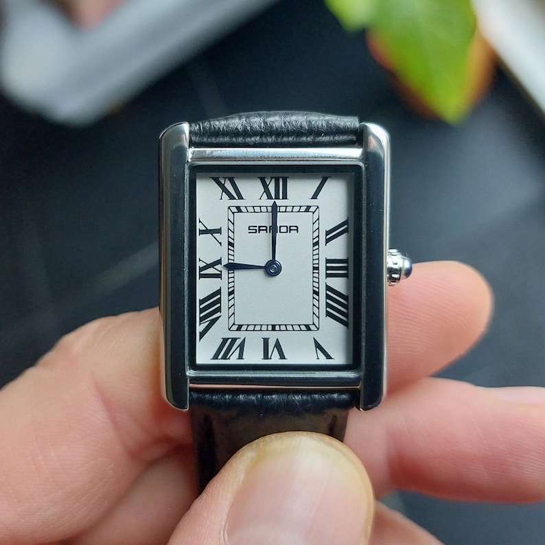 May include: A rectangular wristwatch with a silver-tone case and a black leather band. The watch face is white with black Roman numerals and the brand name "SANDA". The watch hands are blue.