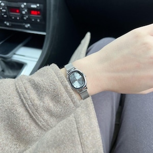 May include: A silver wristwatch with an oval face and a mesh band. The watch face is a light gray color. The watch is worn on a wrist, with a tan coat sleeve and gray pants visible in the background.