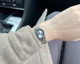 May include: A silver wristwatch with an oval face and a mesh band. The watch face is a light gray color. The watch is worn on a wrist, with a tan coat sleeve and gray pants visible in the background.