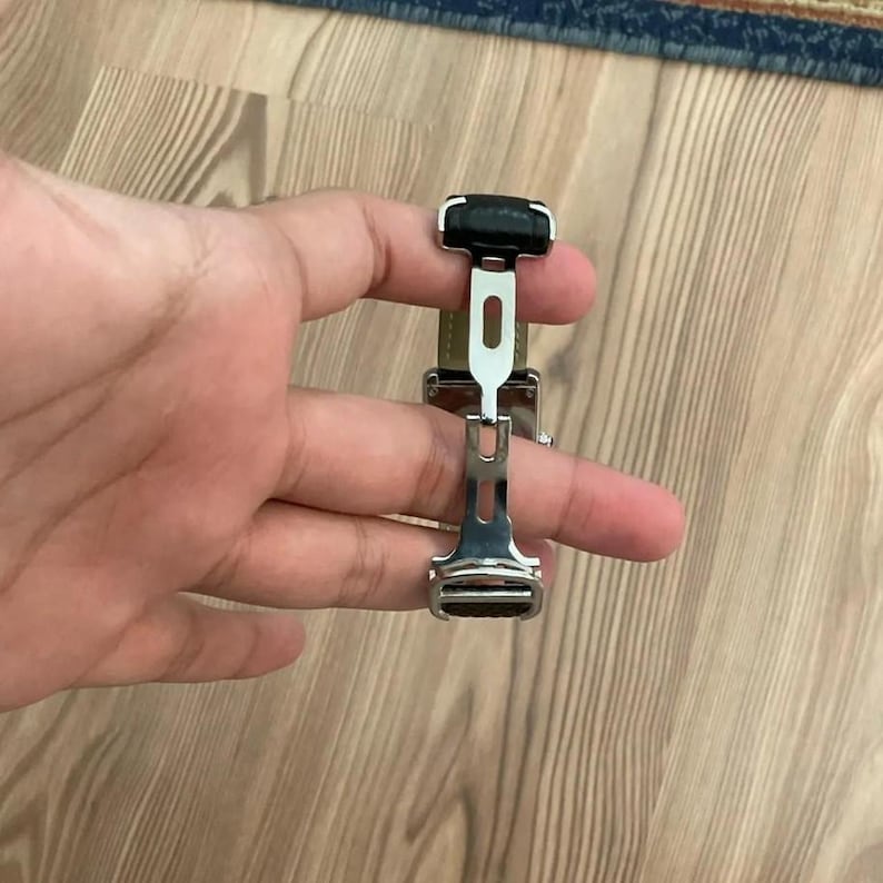 May include: A silver-colored watch clasp with a black leather accent. The clasp is open, revealing a light green leather strap. The design is modern and sleek, suitable for various watch styles.