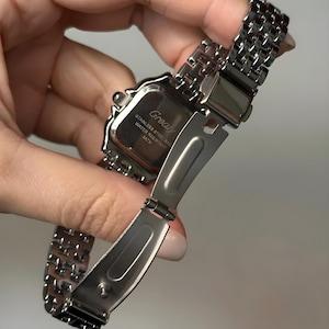 May include: A silver-toned stainless steel watch with a rectangular face and a linked band. The watch face has the word "Grealy" printed on it. The watch is water-resistant.