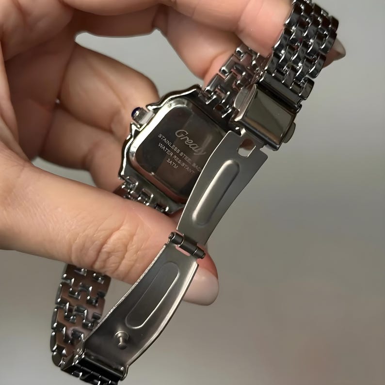May include: A silver-toned stainless steel watch with a rectangular face and a linked band. The watch face has the word "Grealy" printed on it. The watch is water-resistant.