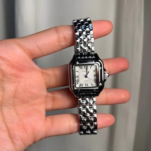 May include: A silver-toned wristwatch with a square face and a linked metal band. The watch face is white with black Roman numerals and hands. The word "QUARTZ" is printed on the watch face.