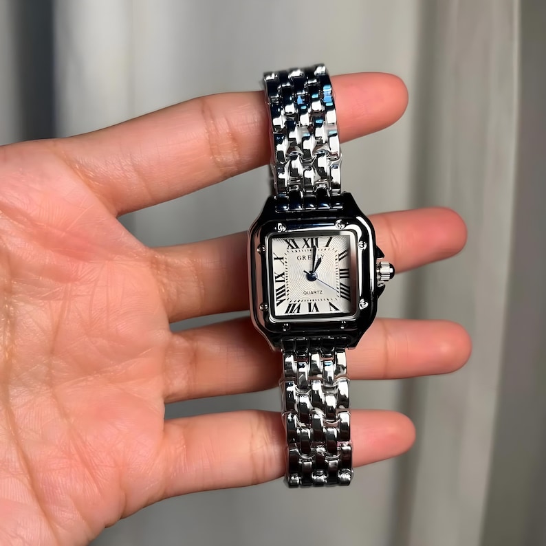 May include: A silver-toned wristwatch with a square face and a linked metal band. The watch face is white with black Roman numerals and hands. The word "QUARTZ" is printed on the watch face.
