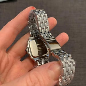 May include: A silver-toned wristwatch with a rectangular face and a linked metal band. The watch is new, still wrapped in plastic. The watch face has the brand name "crealy" printed on it. The watch is water resistant.