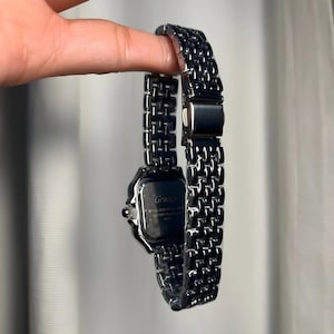 May include: A silver-toned watch with a linked band and a square watch face. The watch face has a dark background with the brand name "Grealy" printed on it. The watch is held up by a hand.