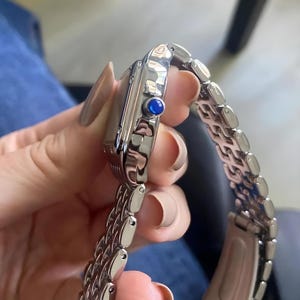 May include: A silver-toned wristwatch with a rectangular face and a linked bracelet. The watch features a blue gemstone on the side. The watch is held by a person.