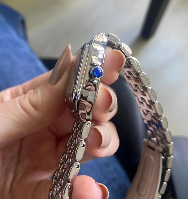 May include: A silver-toned wristwatch with a rectangular face and a linked bracelet. The watch features a blue gemstone on the side. The watch is held by a person.