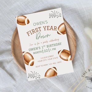 May include: A light beige invitation card with brown football illustrations and text. The text reads "OWEN'S FIRST YEAR DOWN" and invites guests to a first birthday party. The card is on a wooden plate.