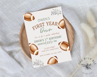 May include: A light beige invitation card with brown football illustrations and text. The text reads "OWEN'S FIRST YEAR DOWN" and invites guests to a first birthday party. The card is on a wooden plate.