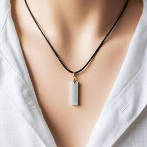 Op de afbeelding: Een rechthoekige hangerketting met een lichtgroene steen. De hanger hangt aan een zwart koord en is bevestigd met een goudkleurige sluiting. De ketting wordt gedragen over een wit overhemd.