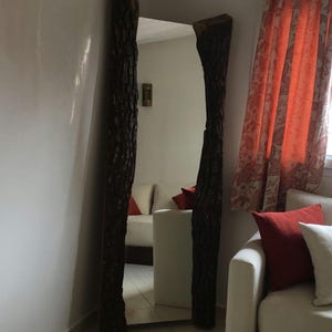 Pode incluir: Espelho de corpo inteiro com uma superfície refletora retangular emoldurada por dois suportes de madeira castanha escura e texturizada. Os suportes assemelham-se a troncos de árvores, adicionando uma estética natural e rústica. O espelho está contra uma parede branca.