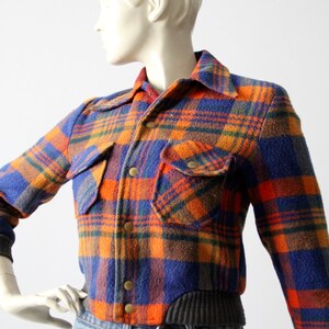 May include: A blue, orange, and green plaid flannel shirt jacket with a ribbed knit bottom band. The jacket has two flap pockets and a button front closure.