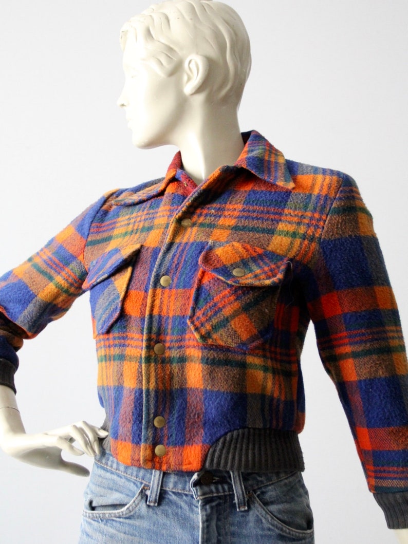 May include: A blue, orange, and green plaid flannel shirt jacket with a ribbed knit bottom band. The jacket has two flap pockets and a button front closure.