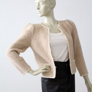 May include: A cream-colored cardigan with a button-up front and puff sleeves. The cardigan has a subtle textured knit and a white trim around the edges.