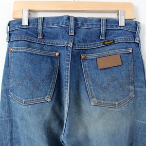 K&ouml;nnte beinhalten: Eine blaue Jeanshose mit einem braunen Leder-Patch auf der Ges&auml;&szlig;tasche, auf dem "Wrangler" steht.