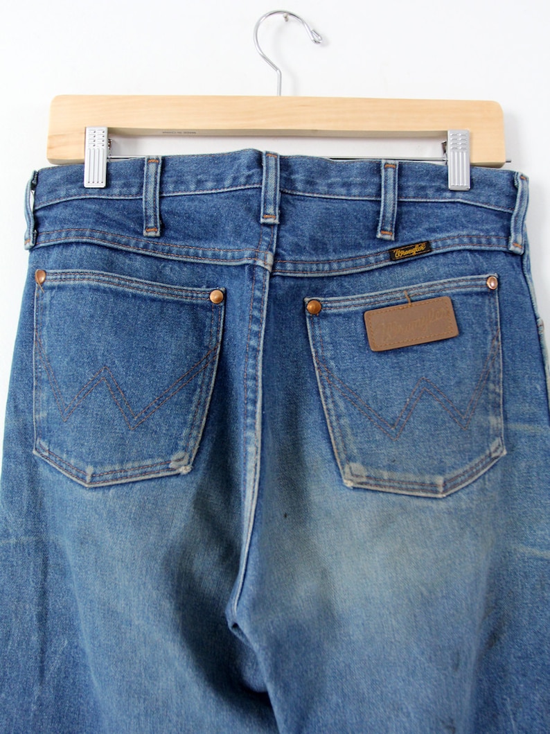 K&ouml;nnte beinhalten: Eine blaue Jeanshose mit einem braunen Leder-Patch auf der Ges&auml;&szlig;tasche, auf dem "Wrangler" steht.