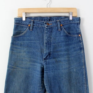 K&ouml;nnte beinhalten: Eine blaue Jeans aus Denim, die an einem Holzb&uuml;gel h&auml;ngt. Die Jeans hat eine hohe Taille und ein gerades Bein.