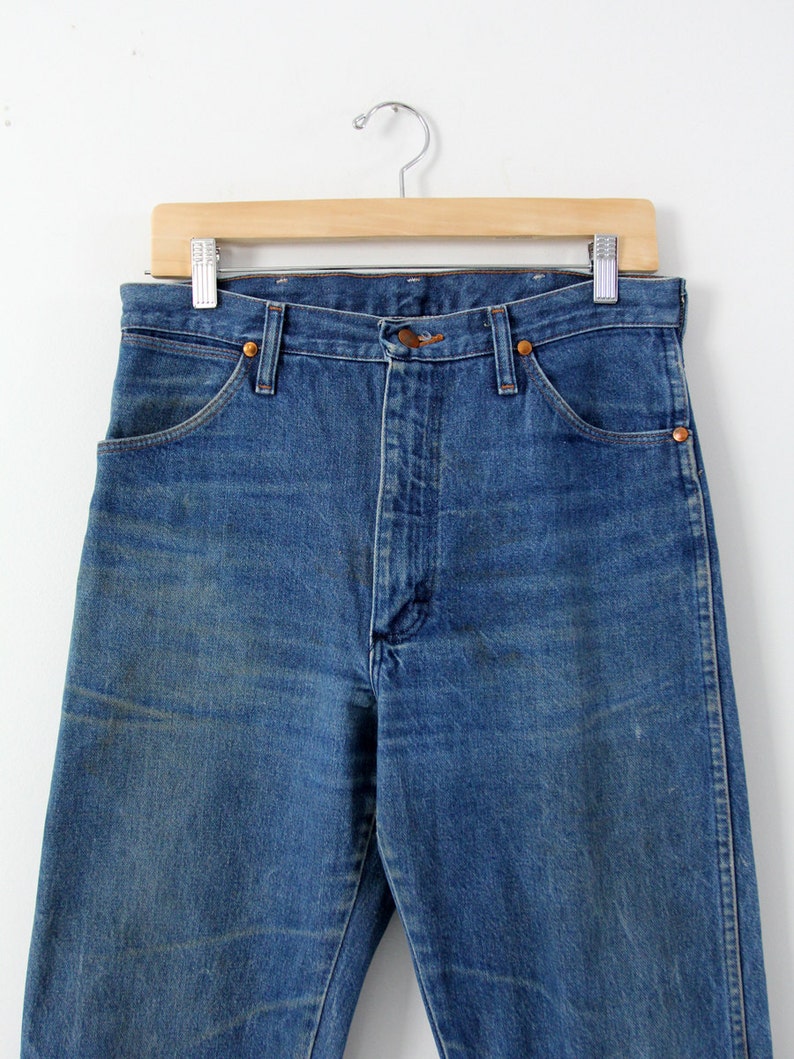 K&ouml;nnte beinhalten: Eine blaue Jeans aus Denim, die an einem Holzb&uuml;gel h&auml;ngt. Die Jeans hat eine hohe Taille und ein gerades Bein.