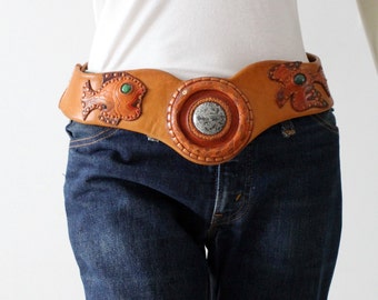 May include: A brown leather belt with a large circular buckle featuring a carved stone design. The belt is decorated with two fish motifs and green stones.