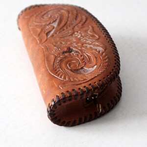 May include: A brown leather clutch purse with intricate floral tooling and a stitched edge. The purse has a zipper closure and a metal ring.