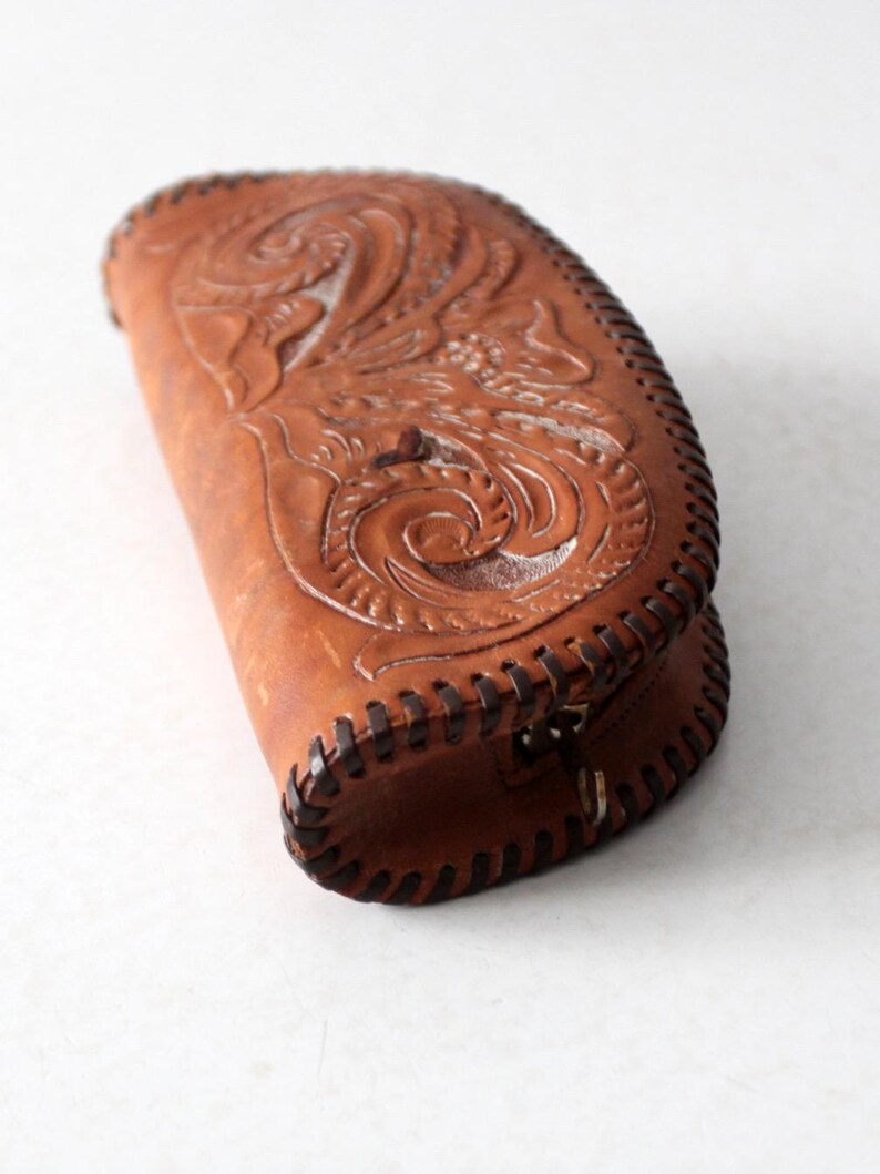 May include: A brown leather clutch purse with intricate floral tooling and a stitched edge. The purse has a zipper closure and a metal ring.