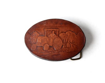 Puede incluir: Hebilla de cinturón ovalada de cuero marrón con diseño de tractor. La hebilla presenta una imagen en relieve detallada de un tractor con un conductor, sobre un paisaje rural. La hebilla tiene un lazo de metal color latón.