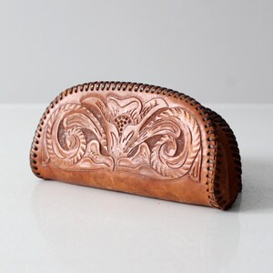 May include: A brown leather clutch purse with a floral design embossed into the leather. The purse has a curved shape and a stitched edge.